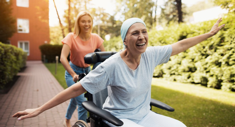 A patient undergoing chemotherapy is celebrating being outside whilst being pushed in a wheelchair by her friend