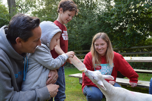 Family of one man, one woman and two boys looking happy and content feeding a baby sheep by milk bottle in a park