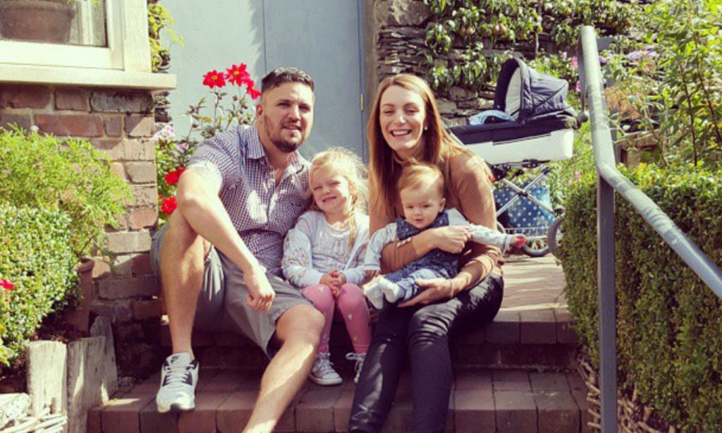 Family of four including two young children sit smiling on their doorstep 