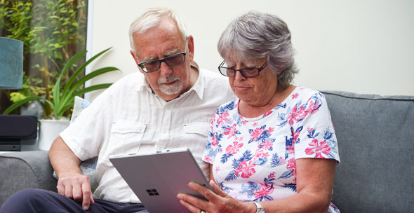 An older couple sit close together on a sofa using a tablet, they look comfortable and deep in thought, perhaps reading about acoustic neuroma.