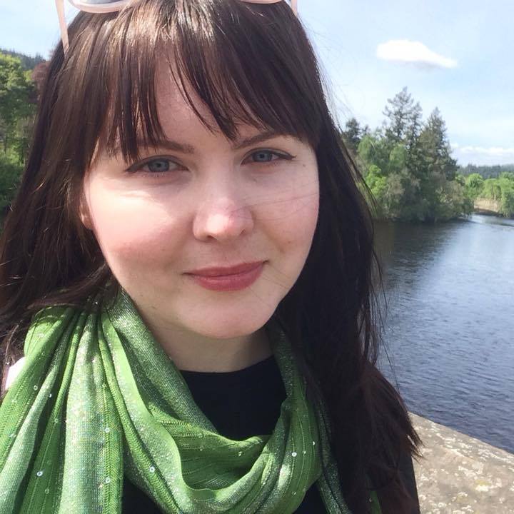 Amy, a 33-year old woman living with a brain tumour, poses for a selfie in front of a beautiful vista full of blue sky, green trees and calm water.