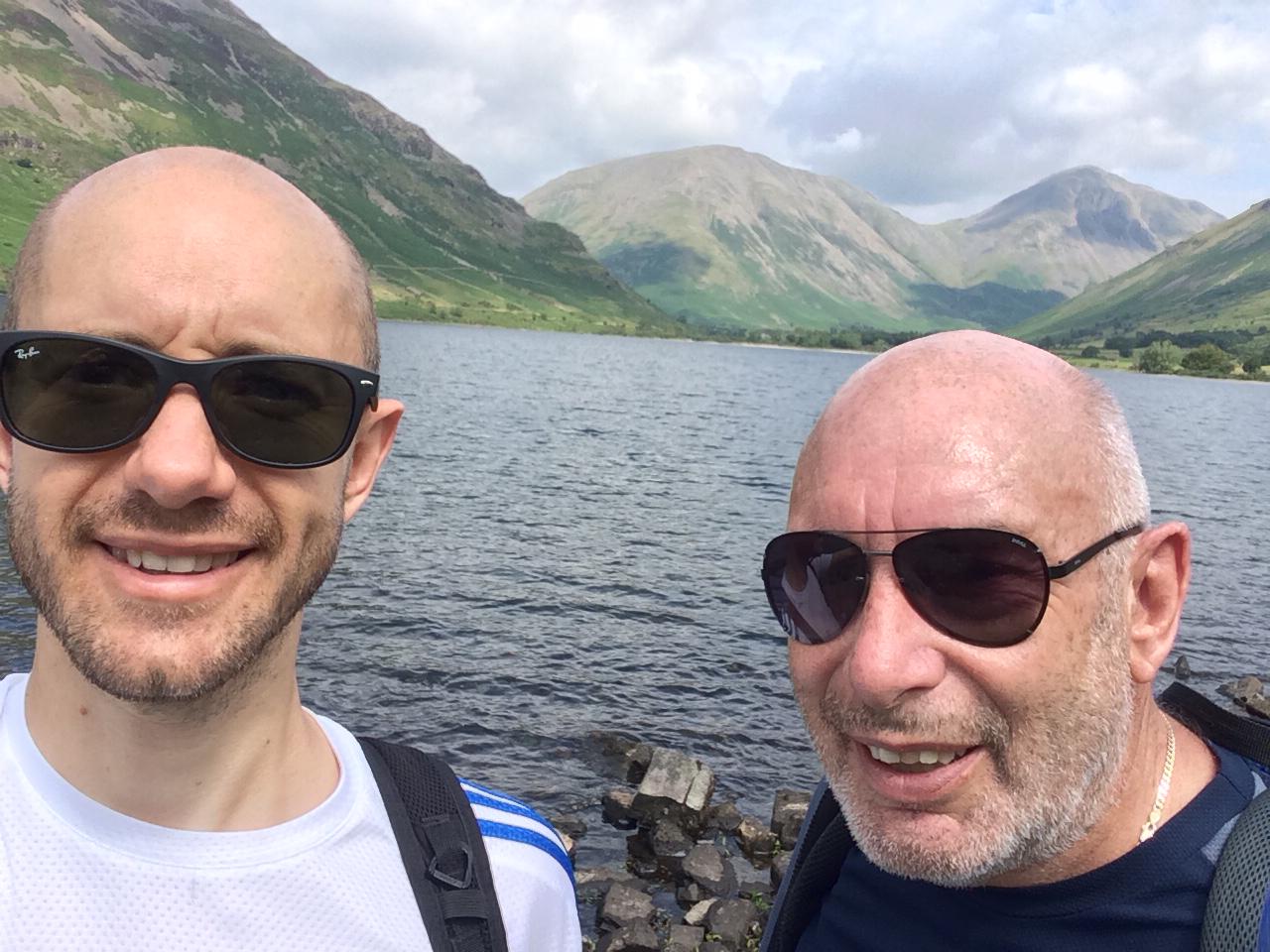 A selfie of two men in front of a lake with mountains in the background.