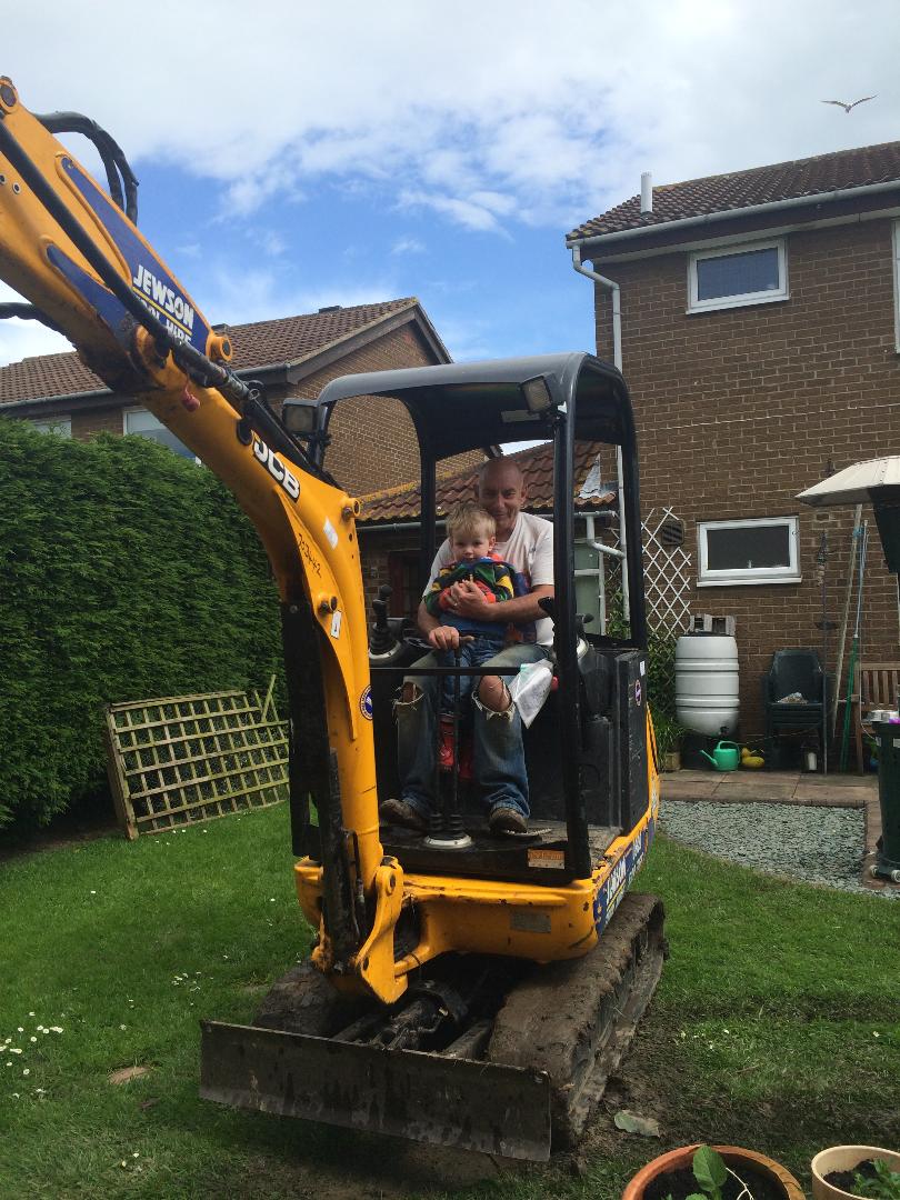 A man with a boy sat on his knee in a yellow digger in a back garden.