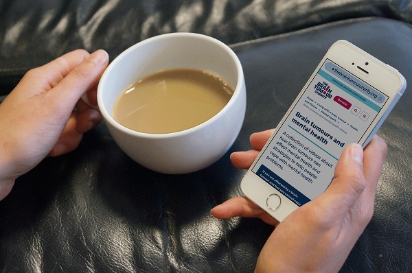 one hand holds a cup of tea while another holds a mobile phone displaying the web page for the free counselling service provided by The Brain Tumour Charity