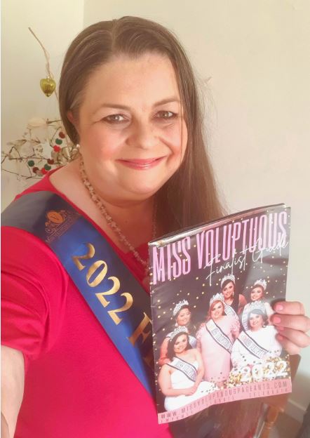 Karen Bucknall wearing a pink dress with a blue pageant sash. Holding a Miss Voluptuous magazine.