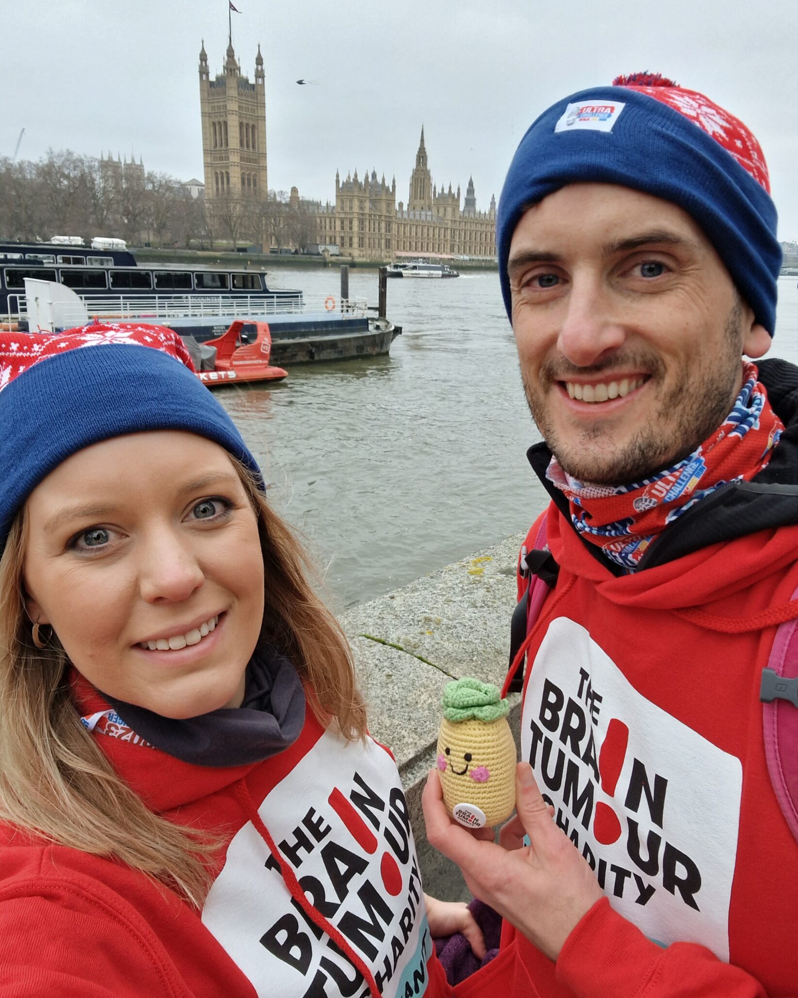 Tom and Lucy wearing The Brain Tumour Charity hoodies standing in front of the Thames in London while on a charity walking challenge