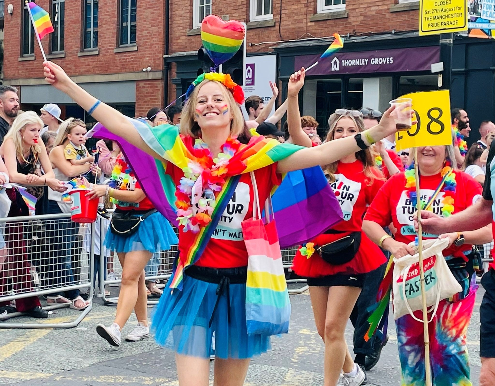 A photo of Laura Nuttall marching at Manchester Pride wearing a The Brain Tumour Charity t-shirt. Laura has a Pride flag draped over her shoulder and she beams as she waves a little Pride flag in one hand and grasps a drink in her other hand.