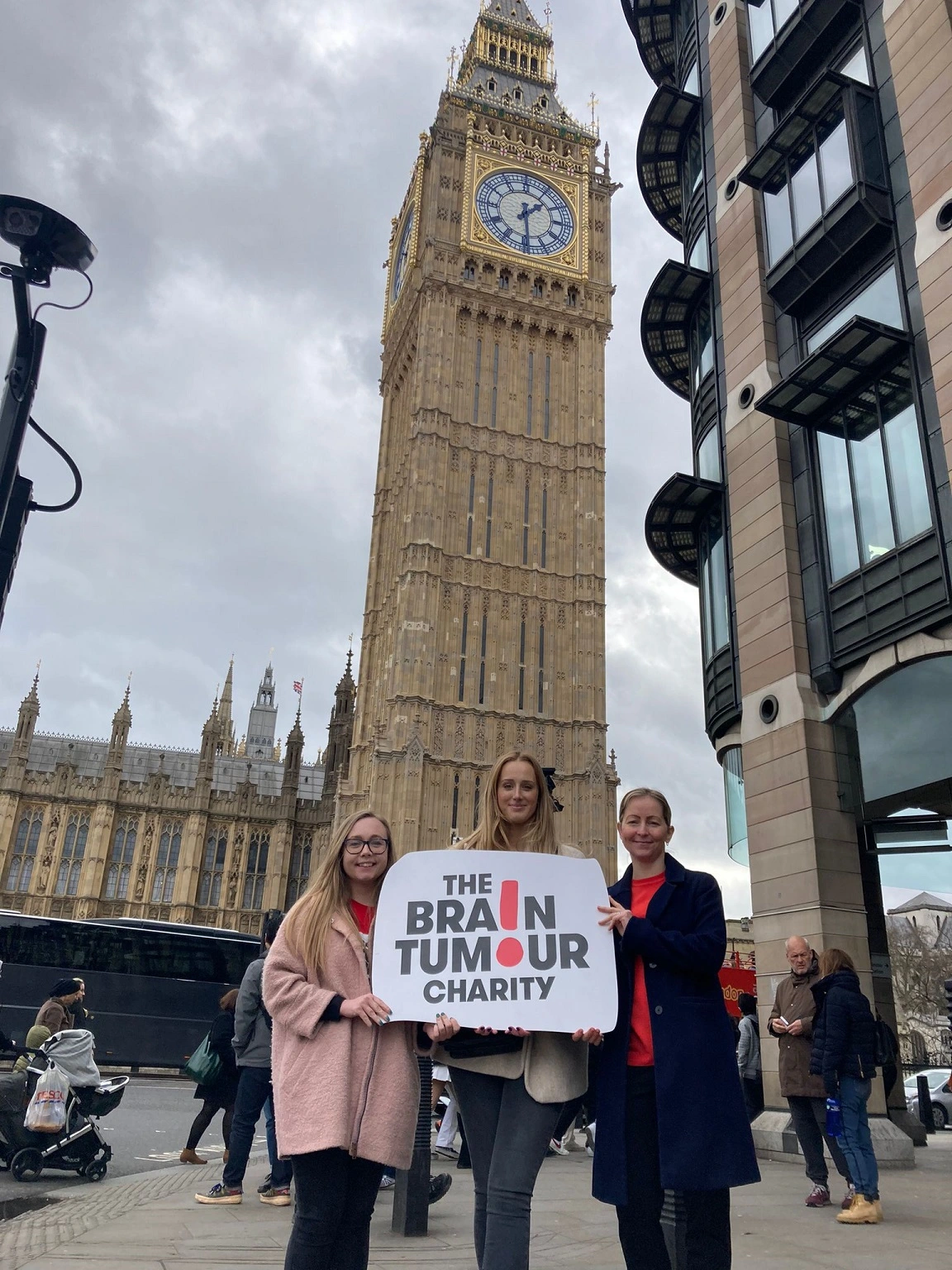 Three volunteers stand outside the Houses of Parliament with a The Brain Tumour Charity sign - we're celebrating them during Volunteers' Week