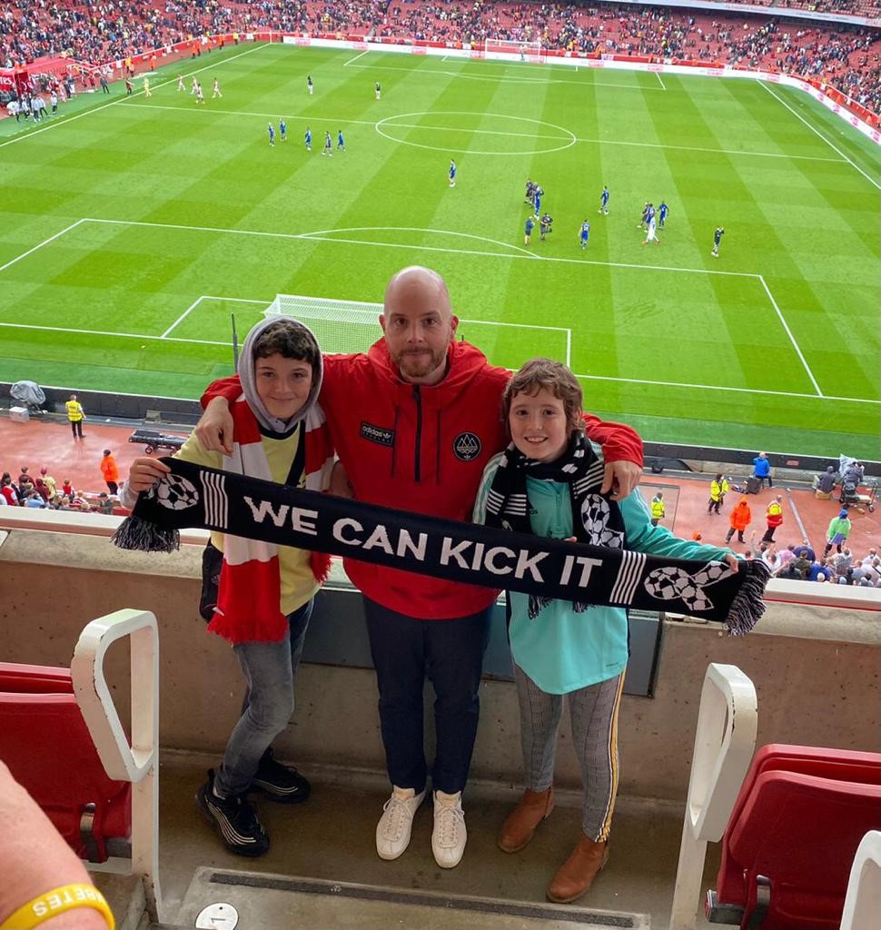 Adrian at a football match with two children, holding a scarf that says We Can Kick It