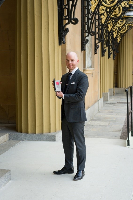 Adrian Conoboy in a suit holding up his MBE award for work done with his football charity We Can Kick It