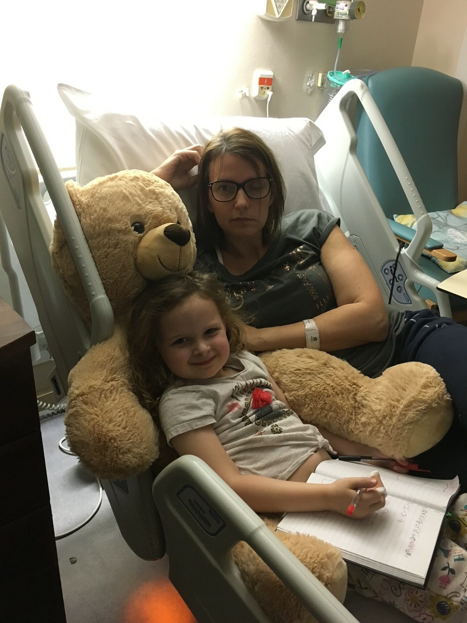 Anya lies in her hospital bed with her daughter resting on a big teddy bear