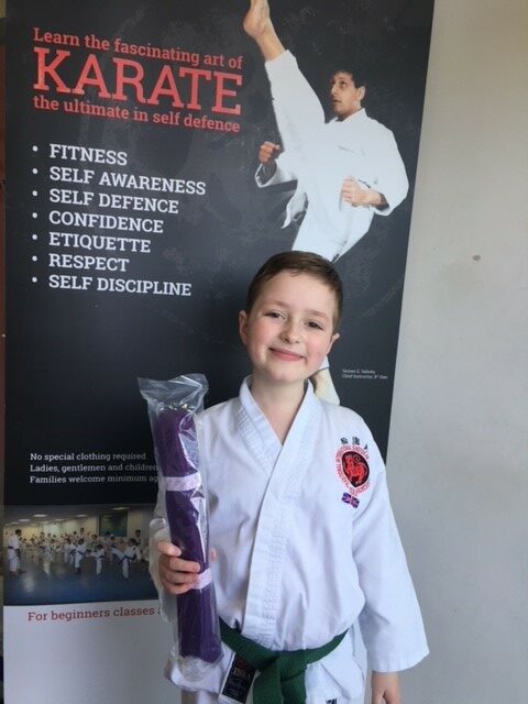 Zac in his white karate kit with green belt., holding his newly awarded purple belt.  He doesn't let his optic nerve glioma diagnosis hold him back.