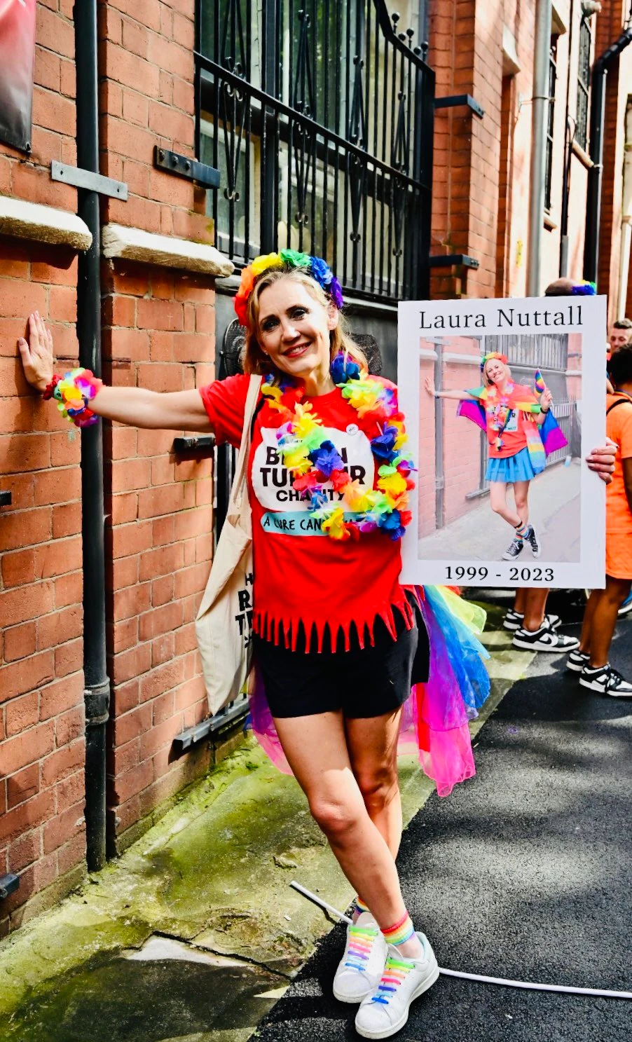 Nicola Nuttall attending PRIDE this year in memory of Laura Nuttall. She leans against a wall in the exact pose that Laura displays in the picture Nicola is holding