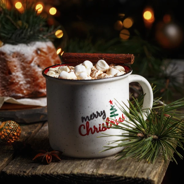 a hot chocolate with marshmallows sits on a wooden table with Christmas lights in the background