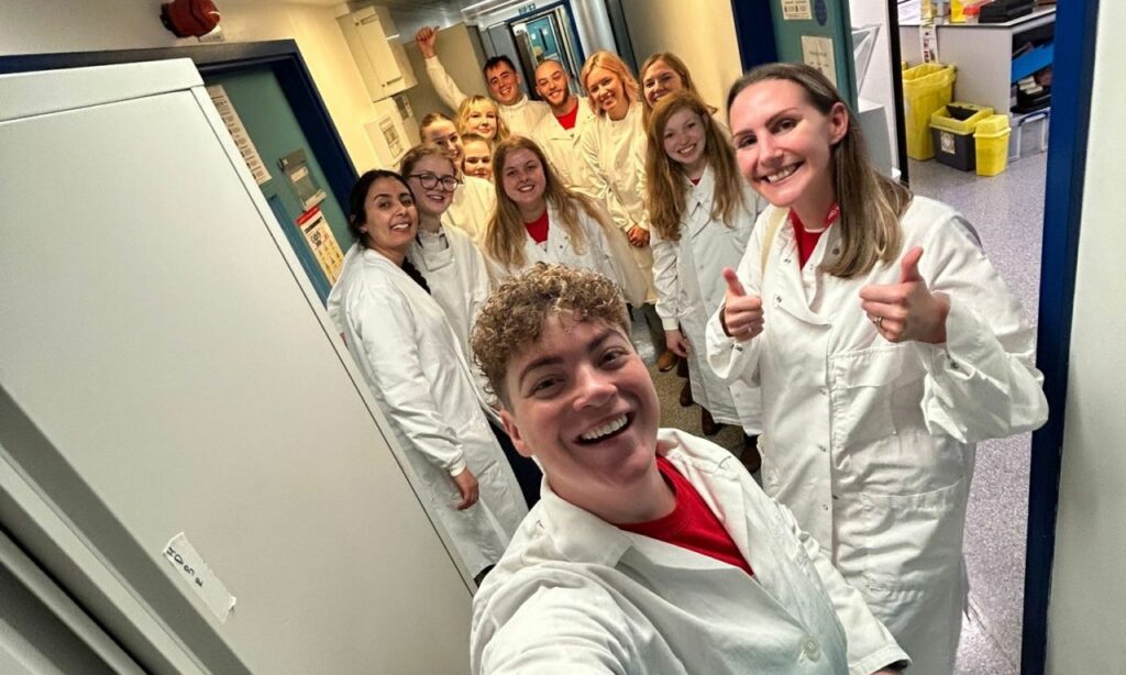 A group of Young Ambassadors in white lab coats take a selfie in a corridor or a laboratory