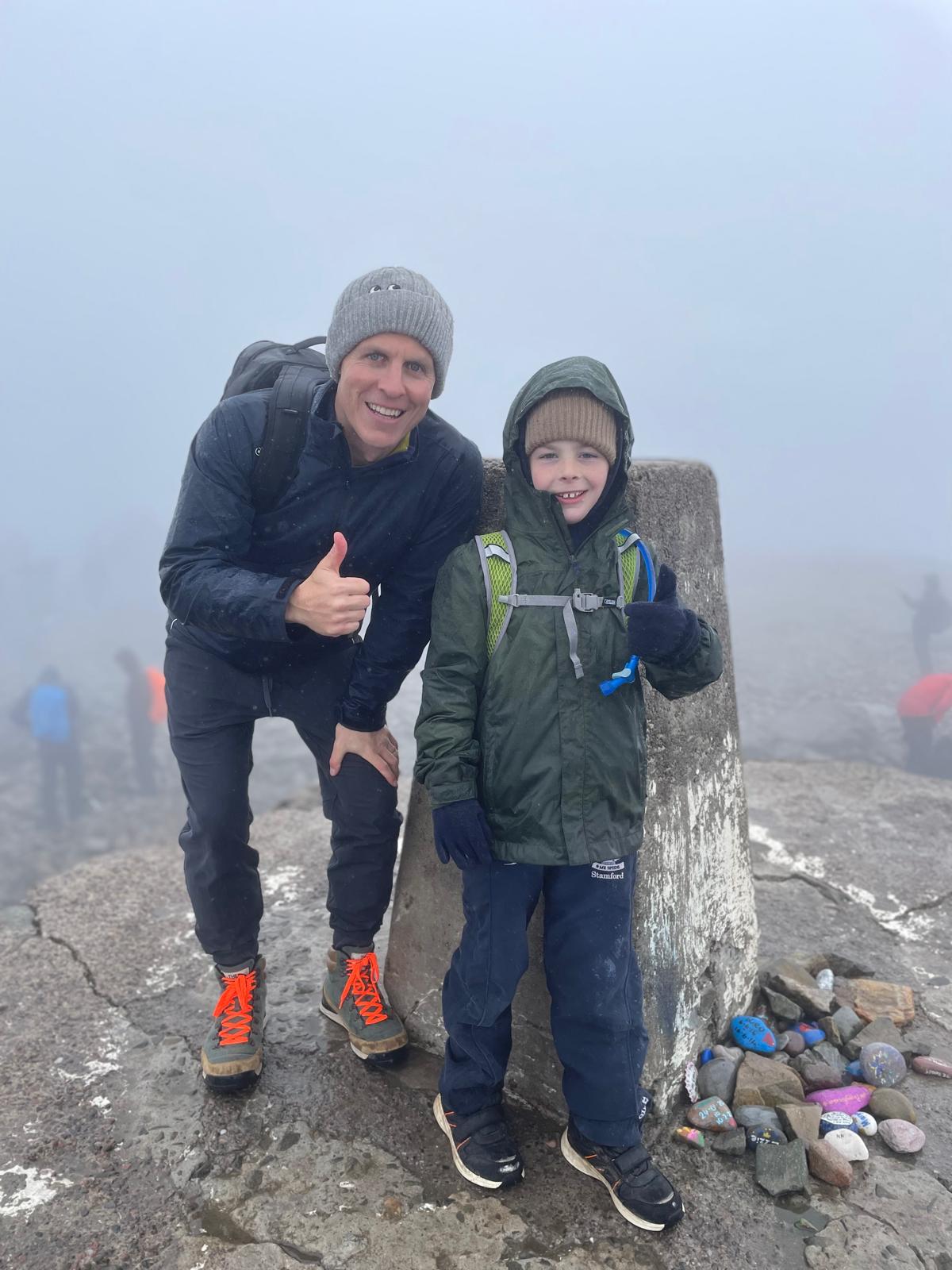 Arthur and his dad stand at the peak of a fell smiling at the camera with their thumbs up.