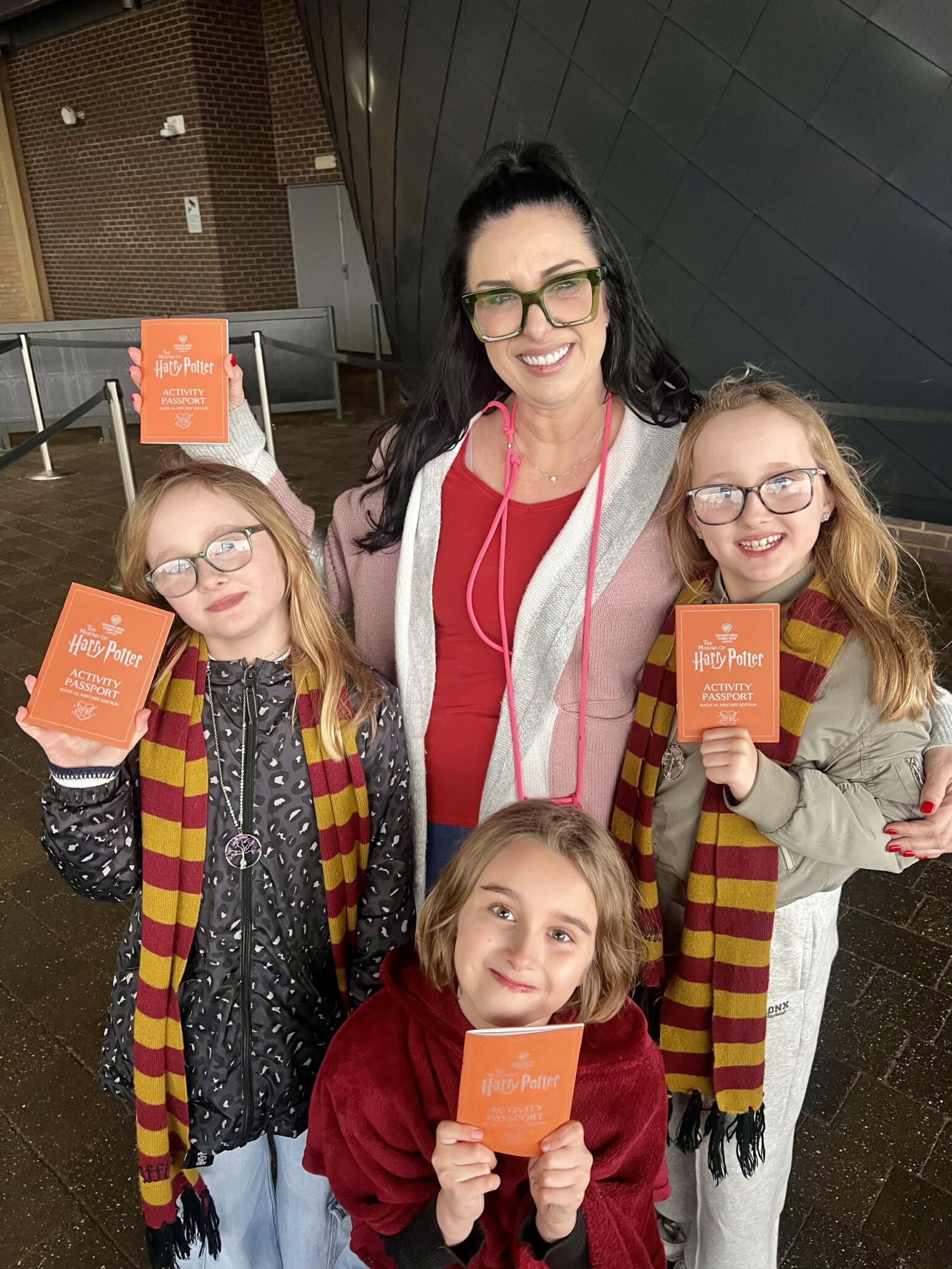 Lisa Connell with three children outside Warner Bros Studio Tour: The Making of Harry Potter. Two are wearing yellow and red striped scarves and all of the are holding activity passports