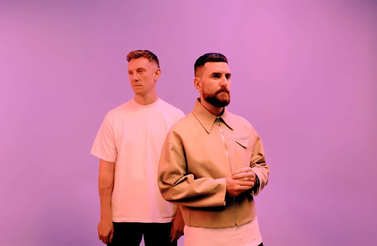 The members of Bicep, the electronic musical duo, stand looking off at different angles with a purple background