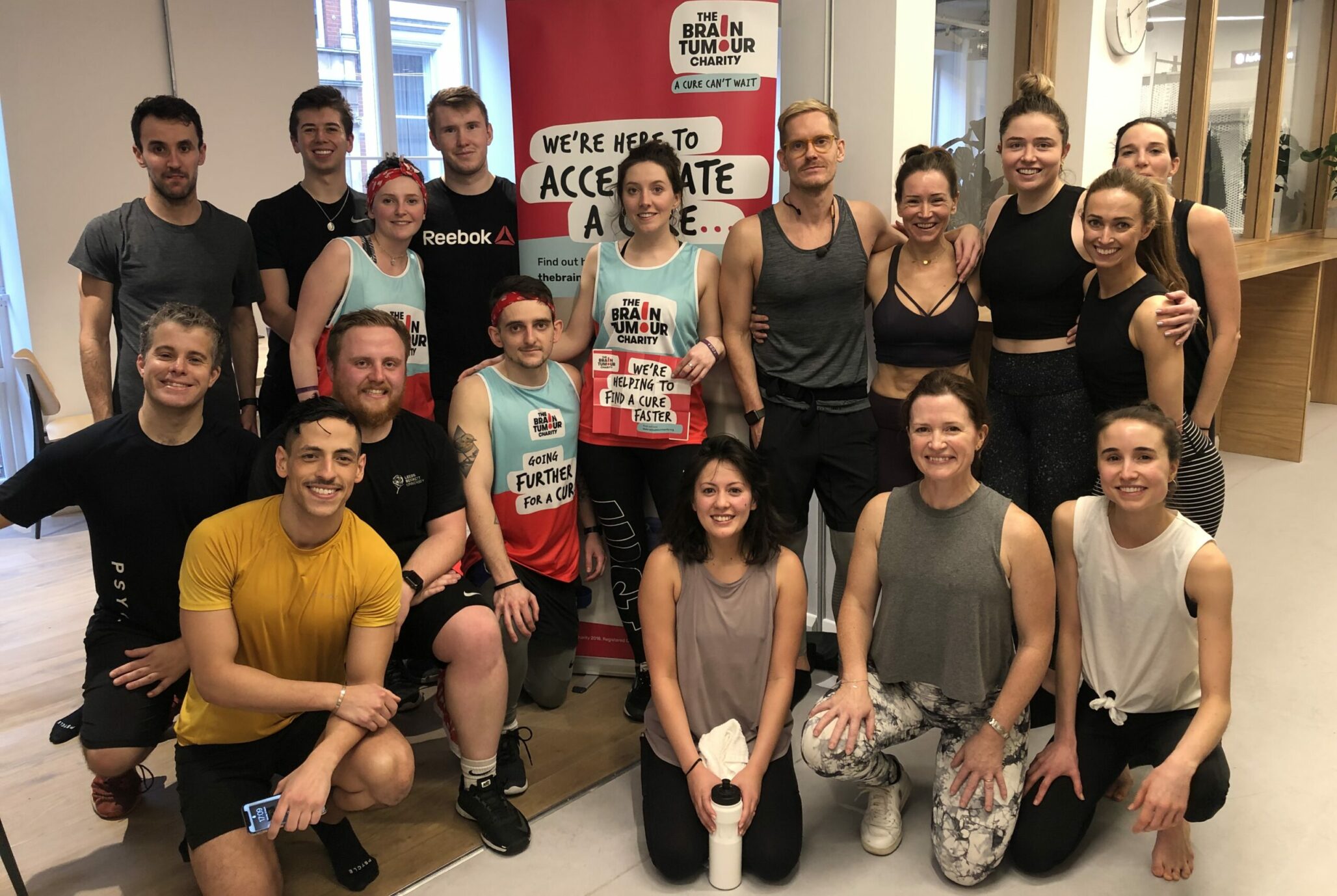 A group of people in fitness clothing huddle around a banner for The Brain Tumour Charity that reads, "We're here to accelerate a cure"
