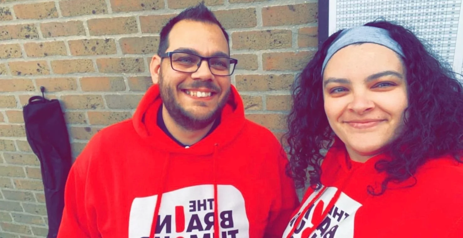 Kasey Brown stands with Liam from The Brain Tumour Charity. They're both wearing red hoodies featuring The Charity's logo