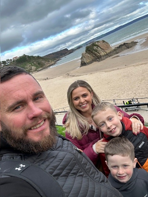 Lewis, his partner, and two boys take a selfie with a beach in the background. Lewis is doing a Twilight Walk Own Walk to raise funds and awareness for brain tumours