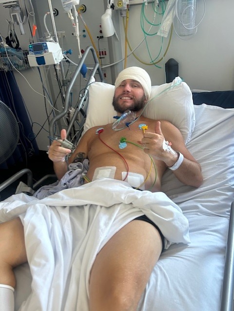 Lewis lies back in his hospital bed and holds two thumbs up