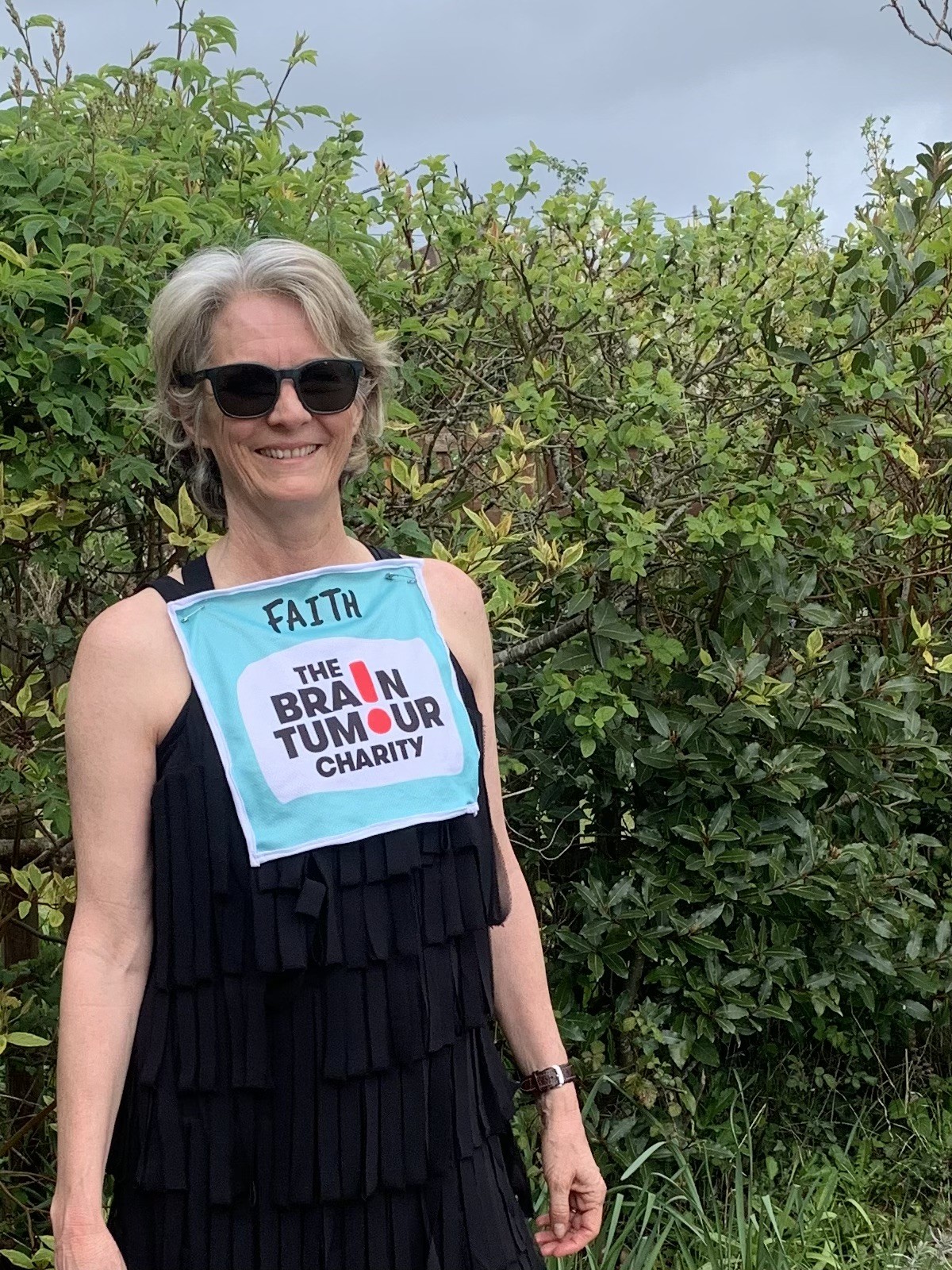 Faith stands wearing a black top with a teal running bib pinned to it, there is a green bush in the background 