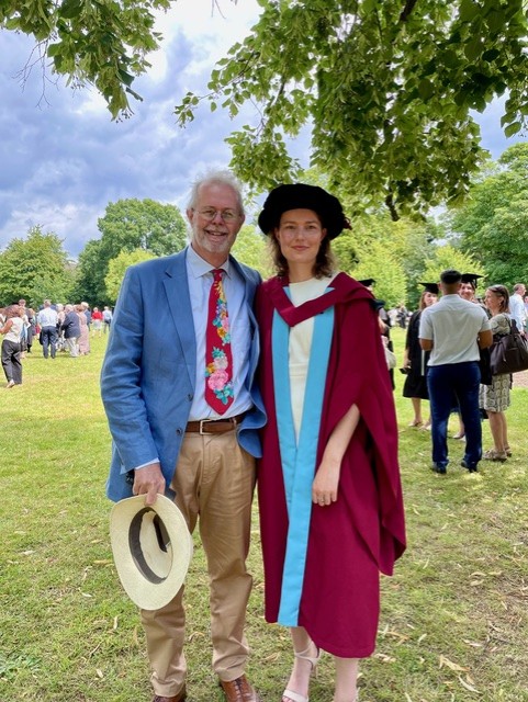 Pippa stands in a burgundy graduation gown with her arm around her father. They stand in a field with tree branches overhead