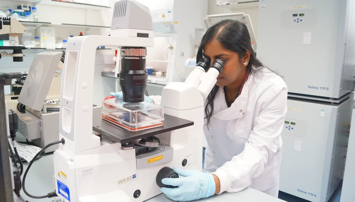 Researcher in a white lab coat looking into a microscope