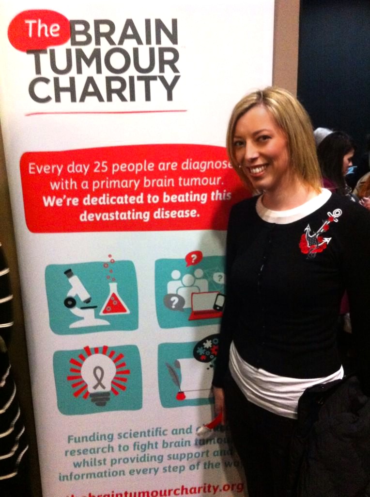 Claire Bullimore, author of A Brain Tumour's Travel Tale, stands next to a banner for The Brain Tumour Charity