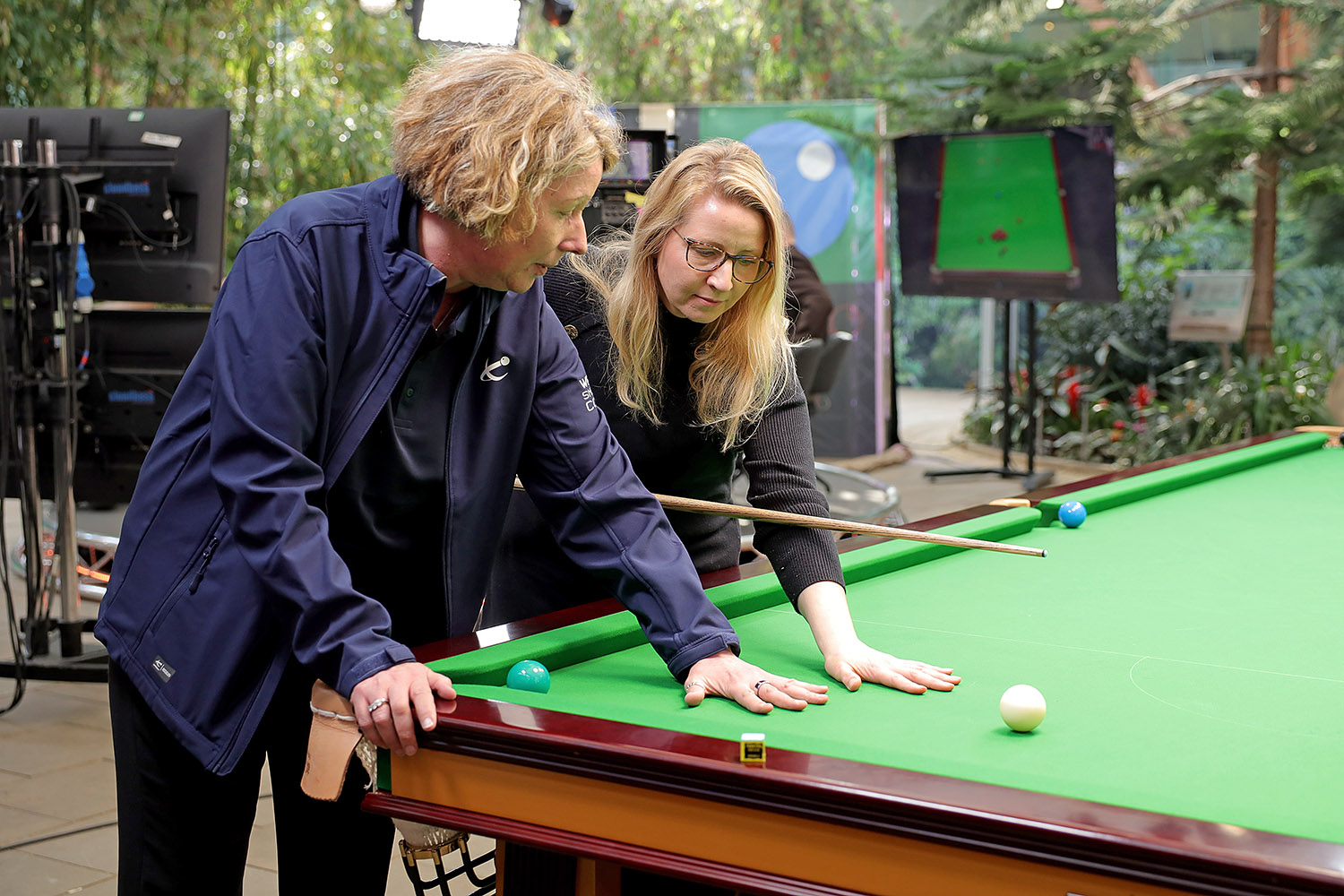 A snooker player teaches another player how to line up a shot on a table as part of a promo shoot for the partnership between the EPSB and The Brain Tumour Charity