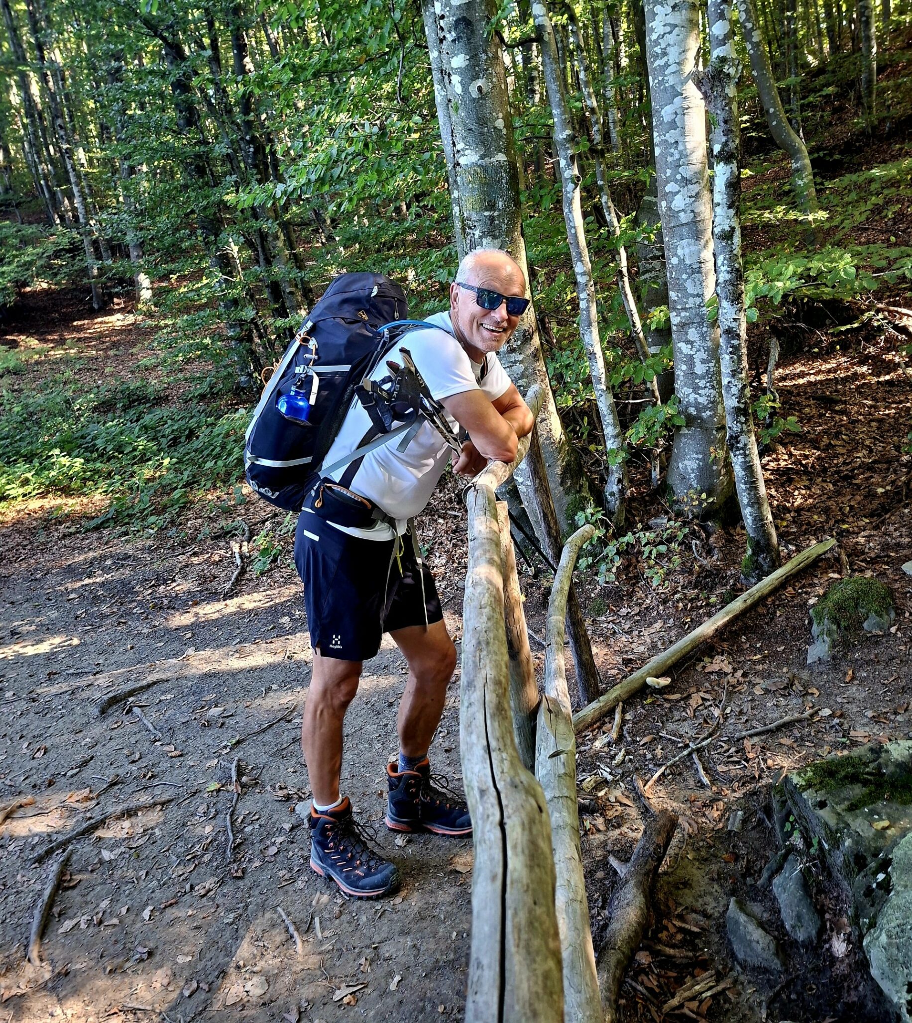 Jeff leans against a wooden fence in a forest, holding walking poles with a backpack on his back