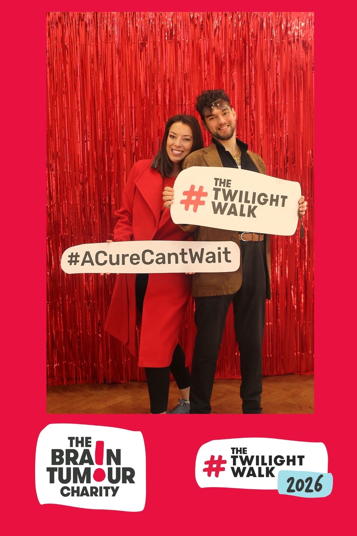 Claire Bullimore stands with a fellow walker at The Twilight Walk. They stand in front of red glittery ribbons and hold sign cards that read #TheTwilightWalk and #ACureCantWait