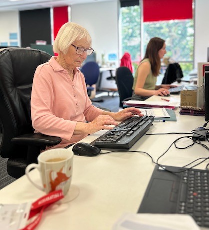 Volunteer Vera is sitting at a desk wearing a pink long sleeved top and using a keyboard