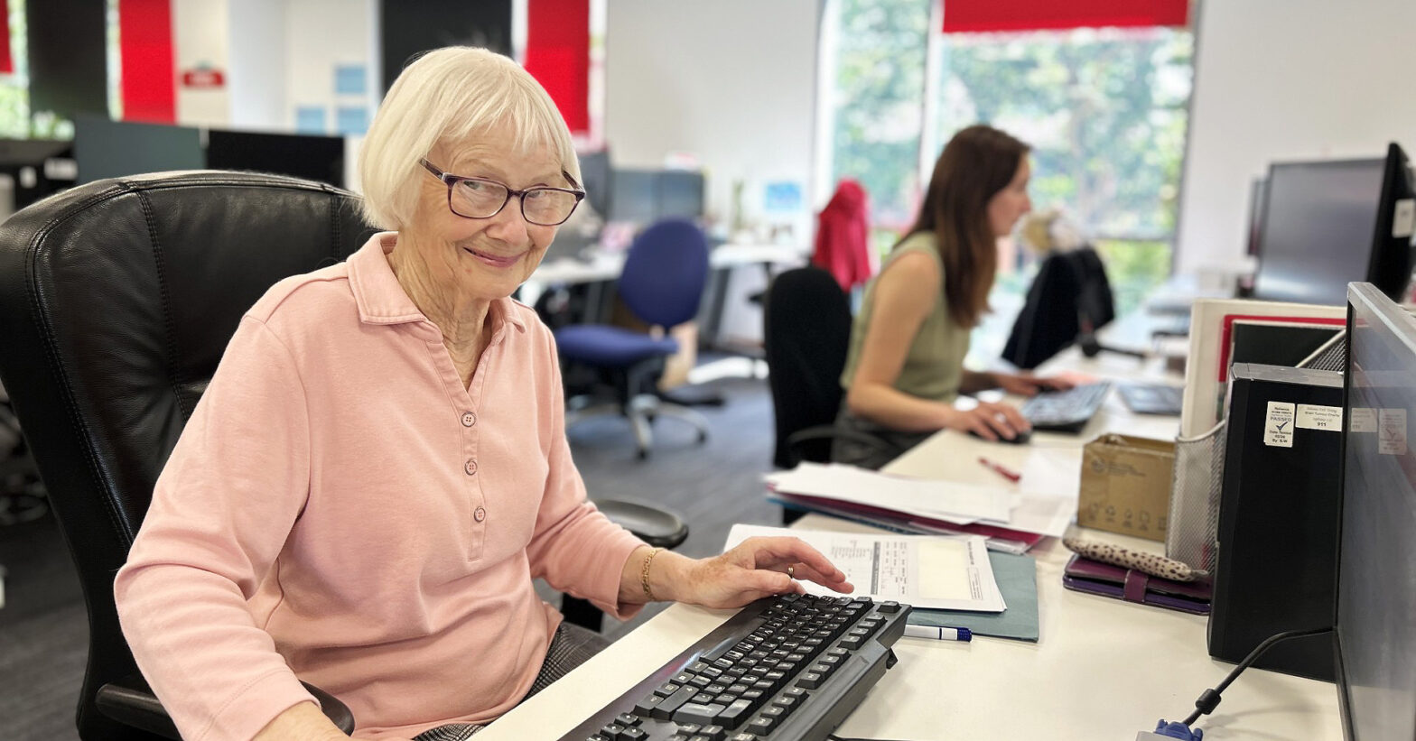 Vera sits in front of a keyboard smiling to camera. She has chin length white hair and glasses