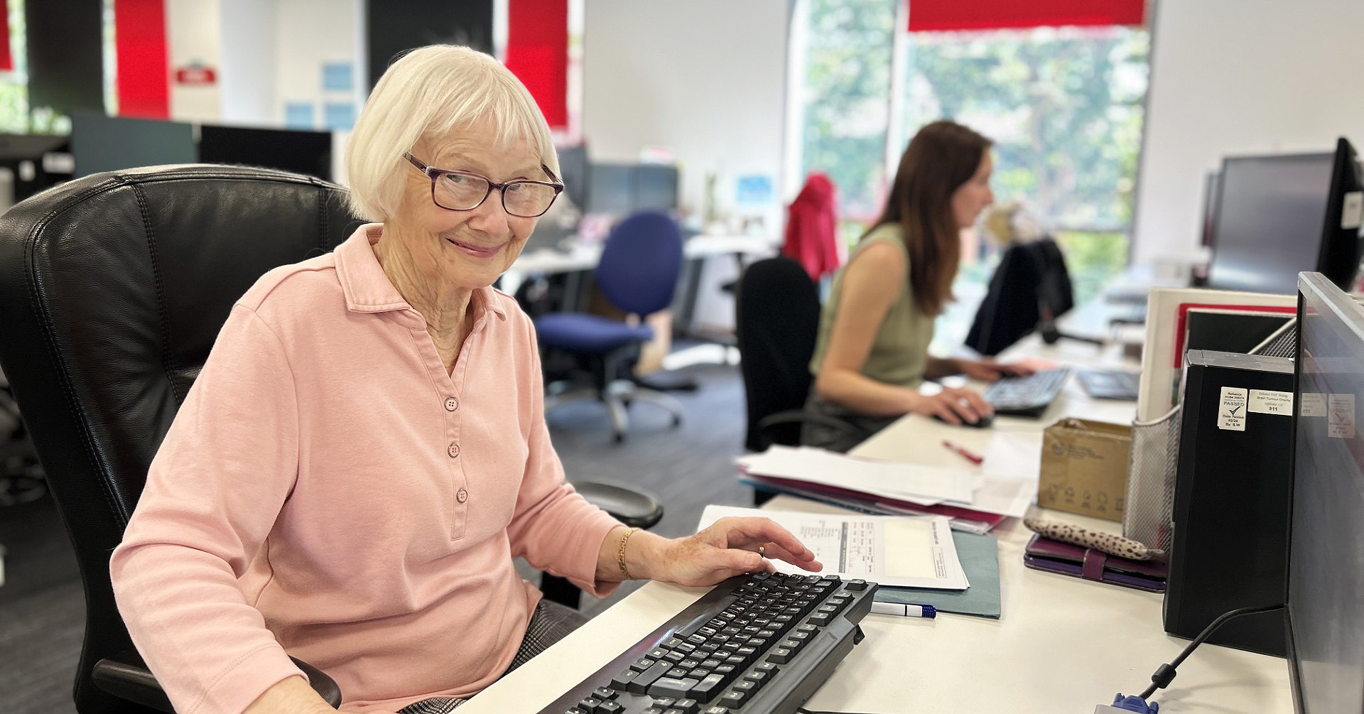 Vera sits in front of a keyboard smiling to camera. She has chin length white hair and glasses