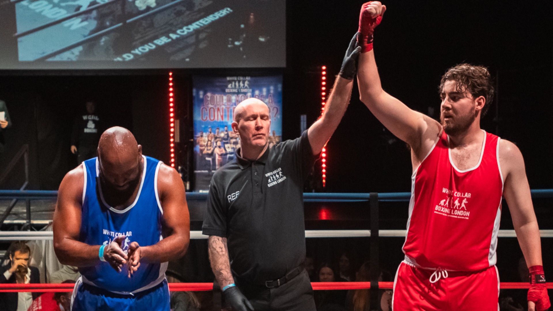 Two boxers stand with the referee in the middle of a boxing ring after hearing the result of the match. One boxer wears a red vest and shorts, the other wears blue. The referee holds the boxer in red's hand in the air.