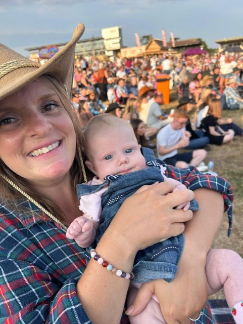 April wears a cowboy hat and sits with her baby on a grass field with a crowd in the background