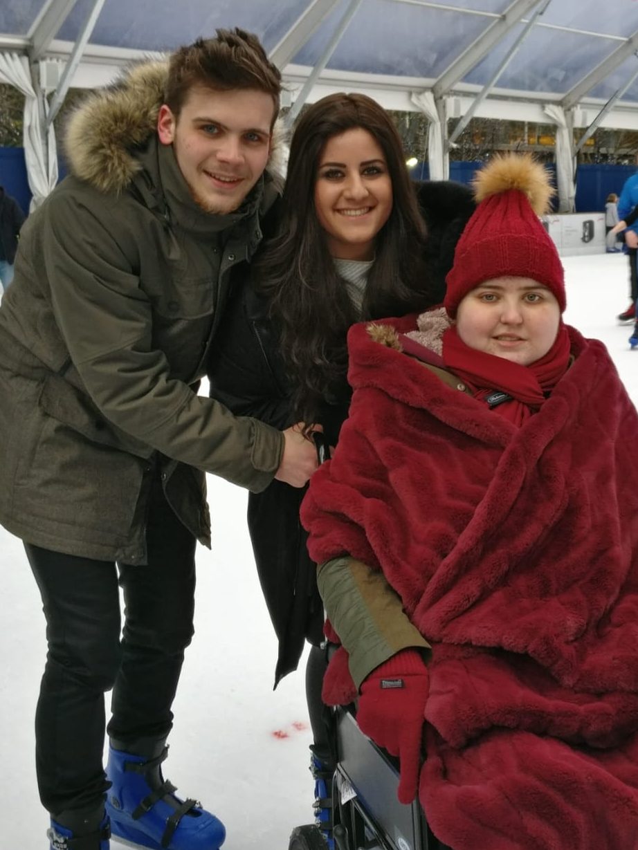 Rachel in a wheelchair covered with a red blanker ice-skating with her brother and Sarah.